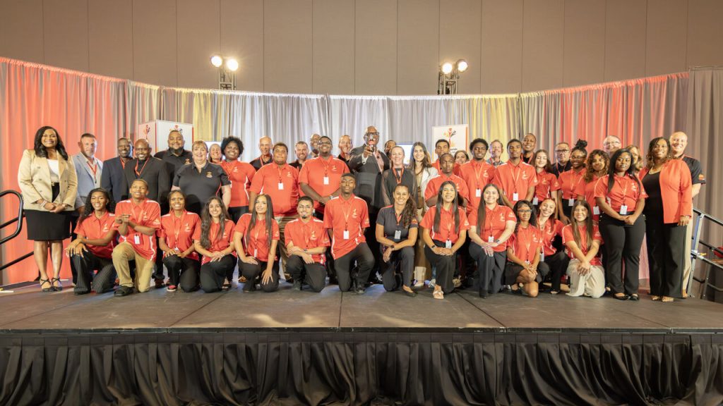 Large group of people on a stage posing for a group photo at an event, many wearing red shirts and name badges.