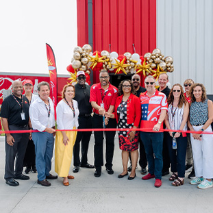 Diverse group of adults at a ribbon-cutting ceremony outside a building with red, white, and gray exterior panels, gold balloons, and a red ribbon as they smile for the celebration.