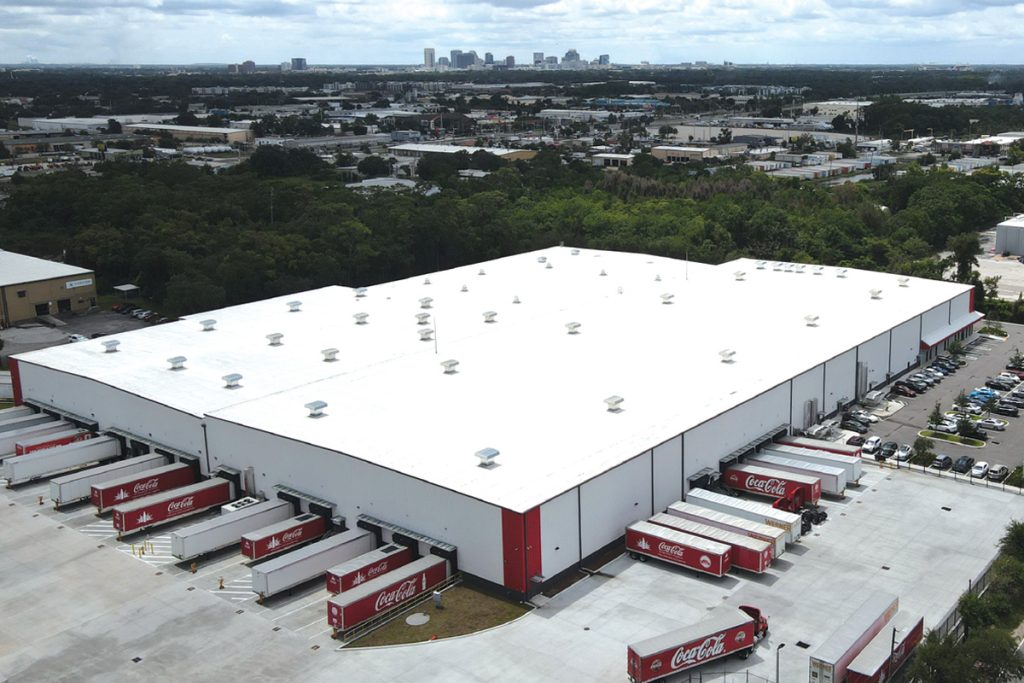 Aerial view of a large white-roofed distribution warehouse with multiple red Coca-Cola trucks parked at loading bays