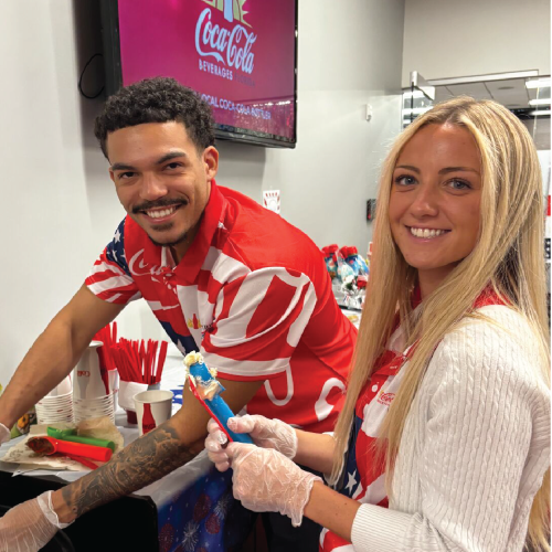 Two people smiling at a Coca-Cola event, preparing blue popsicles at a booth with red-and-white decorations and cups in the background.