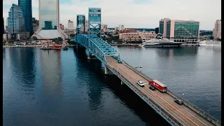 Aerial view of a blue steel bridge crossing a wide river toward a modern city skyline with buildings in the background, cars and a red bus traveling across the bridge.