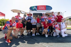 Group of people in red, white, and blue outfits posing in front of a van with a patriotic surfboard sign reading 'Drink in America'; several hold Coca‑Cola cans.