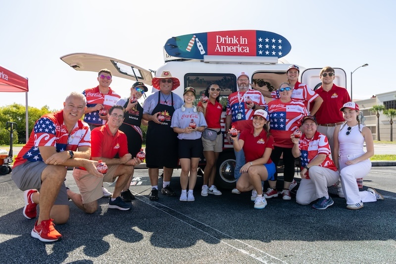 Group of people in red, white, and blue outfits posing in front of a van with a patriotic surfboard sign reading 'Drink in America'; several hold Coca‑Cola cans.