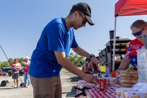 Man in a blue shirt and cap leans over a checkered tablecloth, arranging snacks at an outdoor food stand under a red canopy.