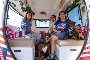 Three women sit in a retro camper van with red, white, and blue decor and two dogs nearby.
