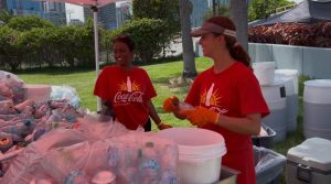Two volunteers in red Coca‑Cola shirts serve drinks at an outdoor event under a canopy, with bags of bottles on the table nearby.