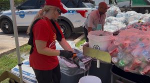 Volunteer in a red shirt and cap, wearing gloves, pouring water from a bottle at a cleanup station with a police SUV in the background.
