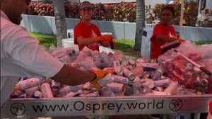 Three volunteers in red shirts and orange gloves sort a pile of crushed aluminum cans at a curbside recycling bin outdoors during a charity event of osprey.world organizer(s).