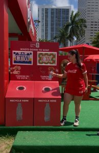 Woman in a red Coca-Cola outfit hands a plastic bottle near red recycling bins at an outdoor promo booth; city high-rises and palm trees in the background.