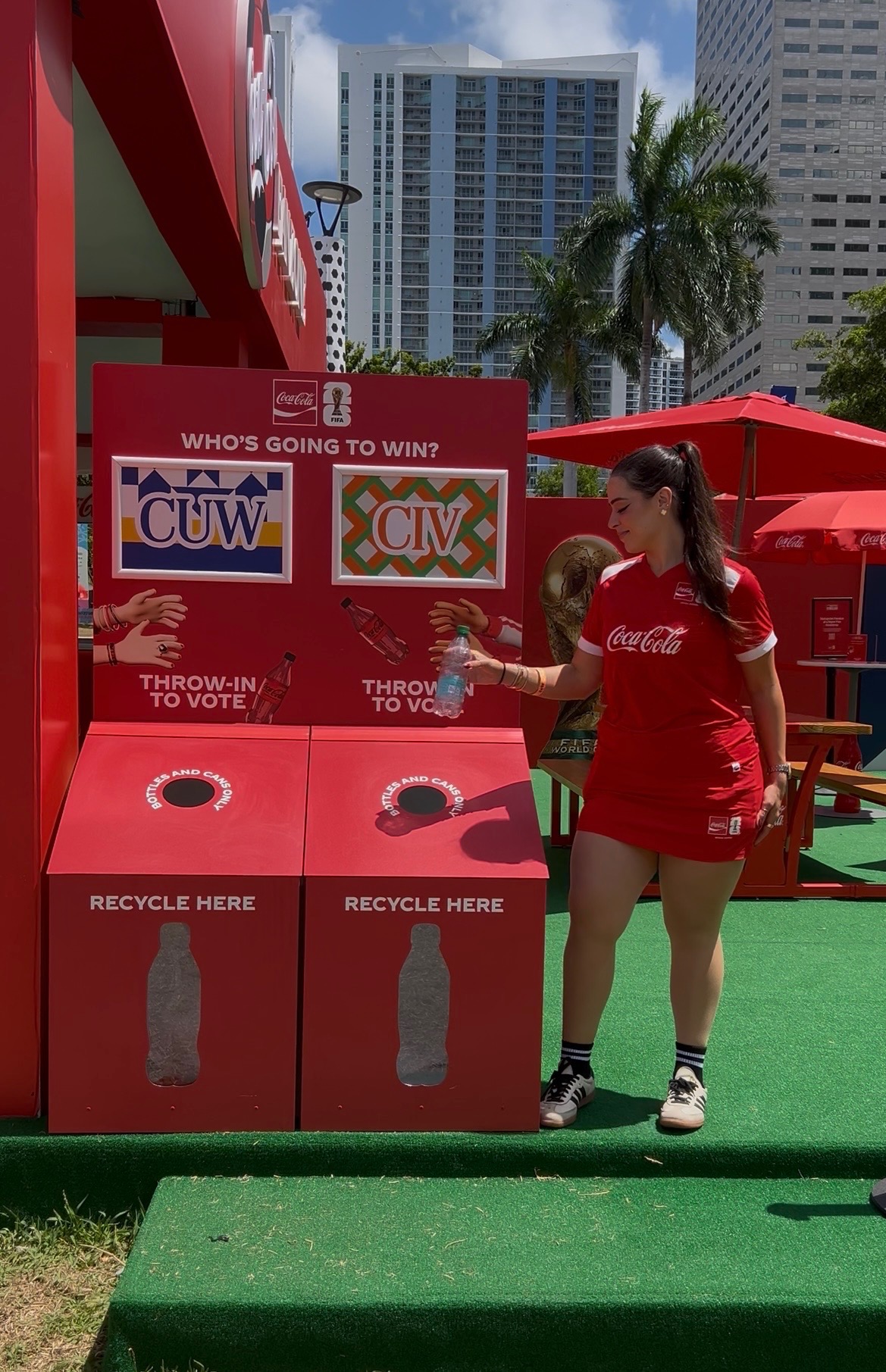 Woman in a red Coca-Cola outfit hands a plastic bottle near red recycling bins at an outdoor promo booth; city high-rises and palm trees in the background.