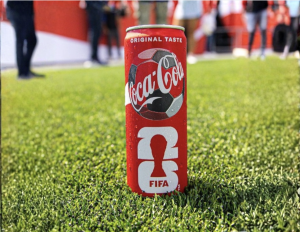 Red Coca‑Cola can on green grass with blurred fans in the background on a stadium field