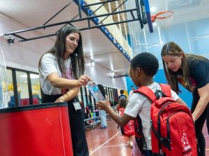 A staff member hands a bottle of water to a smiling child with a red backpack in a gym during a school event.