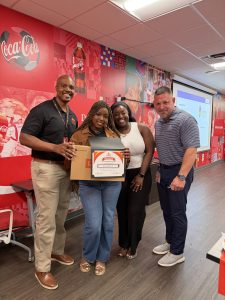 Four people pose for a photo in a Coca‑Cola themed room; the two women hold a certificate and a boxed prize, smiling at the camera.