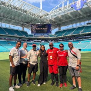 Group of eight people on a stadium grass field posing for a photo with a large Miami banner in the background.