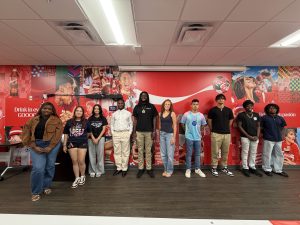 - Coca-Cola Beverages Florida Group of diverse young adults posing for a photo in front of a Coca‑Cola wall mural in a room with ceiling lights.