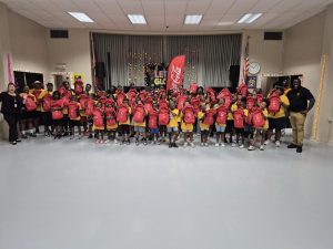 Lincoln Park CC 3 - Coca-Cola Beverages Florida Group of children in yellow shirts holding red backpacks in a decorated school hall, stage backdrop with colorful decorations and Coca-Cola banner.