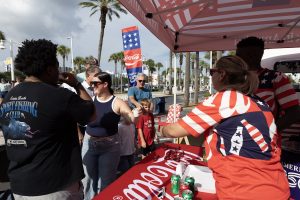 People mingle at a Coca-Cola promotional booth outdoors under a red, flag-themed canopy, palm trees in the background.