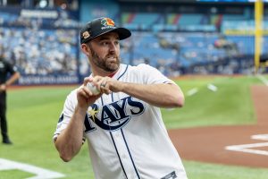 Male baseball pitcher in a Rays uniform, gripping a baseball during a warm-up on the field.
