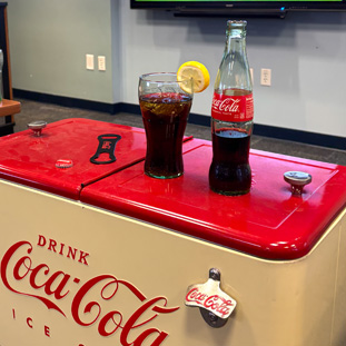 Coca‑Cola cooler with a glass of cola and a lemon wedge on the red lid, plus a Coca‑Cola bottle beside it on top of the cooler.