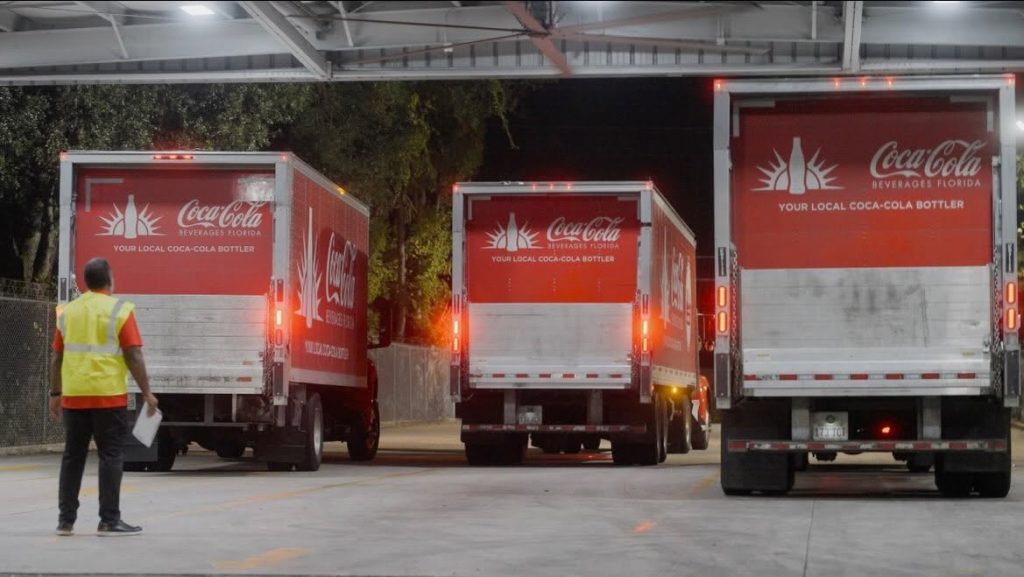 - Coca-Cola Beverages Florida Three Coca-Cola delivery trucks parked in a loading area at night, a worker in a high-visibility vest stands with papers in hand nearby