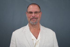 Portrait of a middle-aged man with glasses and a light blazer against a blue-gray backdrop.