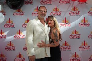 Man in a beige suit and glasses stands with a tattooed woman, posing in front of a Coca-Cola Beverages Florida backdrop.
