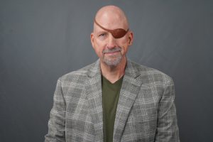 Portrait of a bald man wearing a brown leather eyepatch over one eye, a gray plaid blazer, and a green shirt in a studio backdrop.