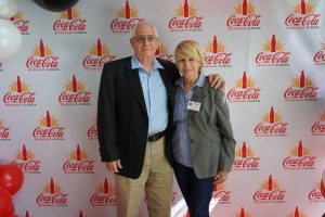 Two smiling adults pose together in front of a Coca-Cola Beverages Florida backdrop at an event, man in a blazer and woman with a name badge.