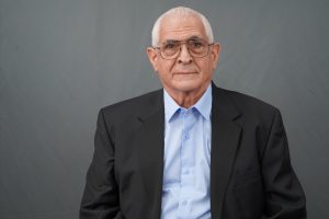 Portrait of an older man with white hair and glasses, wearing a blue dress shirt and dark suit, facing the camera against a gray background.