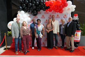 Eight adults pose on a red carpet in front of a Coca‑Cola Beverages Florida backdrop with a black, red, and white balloon arch.