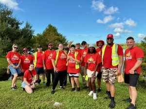 Group of people in red Coca‑Cola shirts posing outdoors on grass, some wearing reflective vests, under a blue sky.