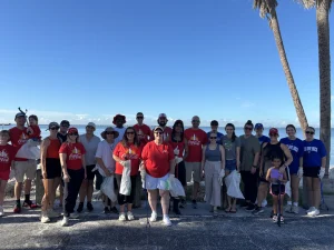 Group of people in red shirts posing together on a sunny beach with palm trees in the background and a blue sky.