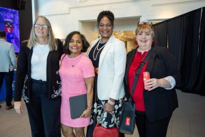 Group of four women posing indoors at an event, smiling for the camera; from left to right: woman with glasses in a black blazer and white top, woman in a pink dress, woman in a white blazer with pearls, and a woman in a red blouse and black blazer holding a red handbag.