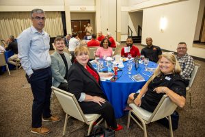 Group of colleagues seated around a round table with a blue tablecloth at a banquet in a conference room, smiling at the camera; one man stands in the foreground left wearing a light blue shirt and brown shoes.