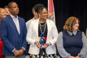 Woman in a white blazer holding a blue glass trophy on a stage, flanked by smiling colleagues and a flag in the background.