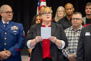 Center woman with blonde hair and glasses holds a paper while speaking, flanked by two men in formal attire at a ceremony.
